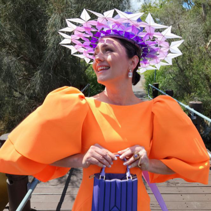 Woman wearing an orange dress and a decorative headpiece with purple and white elements, standing outdoors.