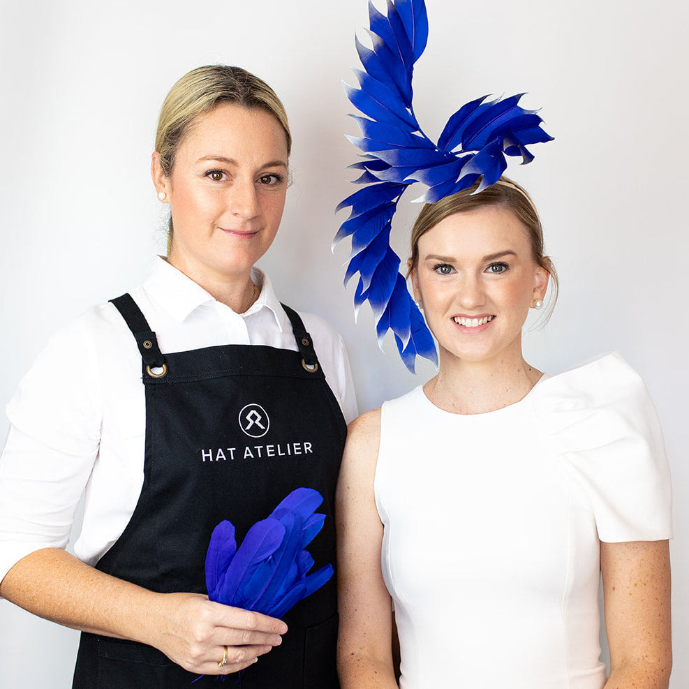 Two women, one wearing a black apron with 'Hat Atelier' branding, holding blue feathered hats against a white background.